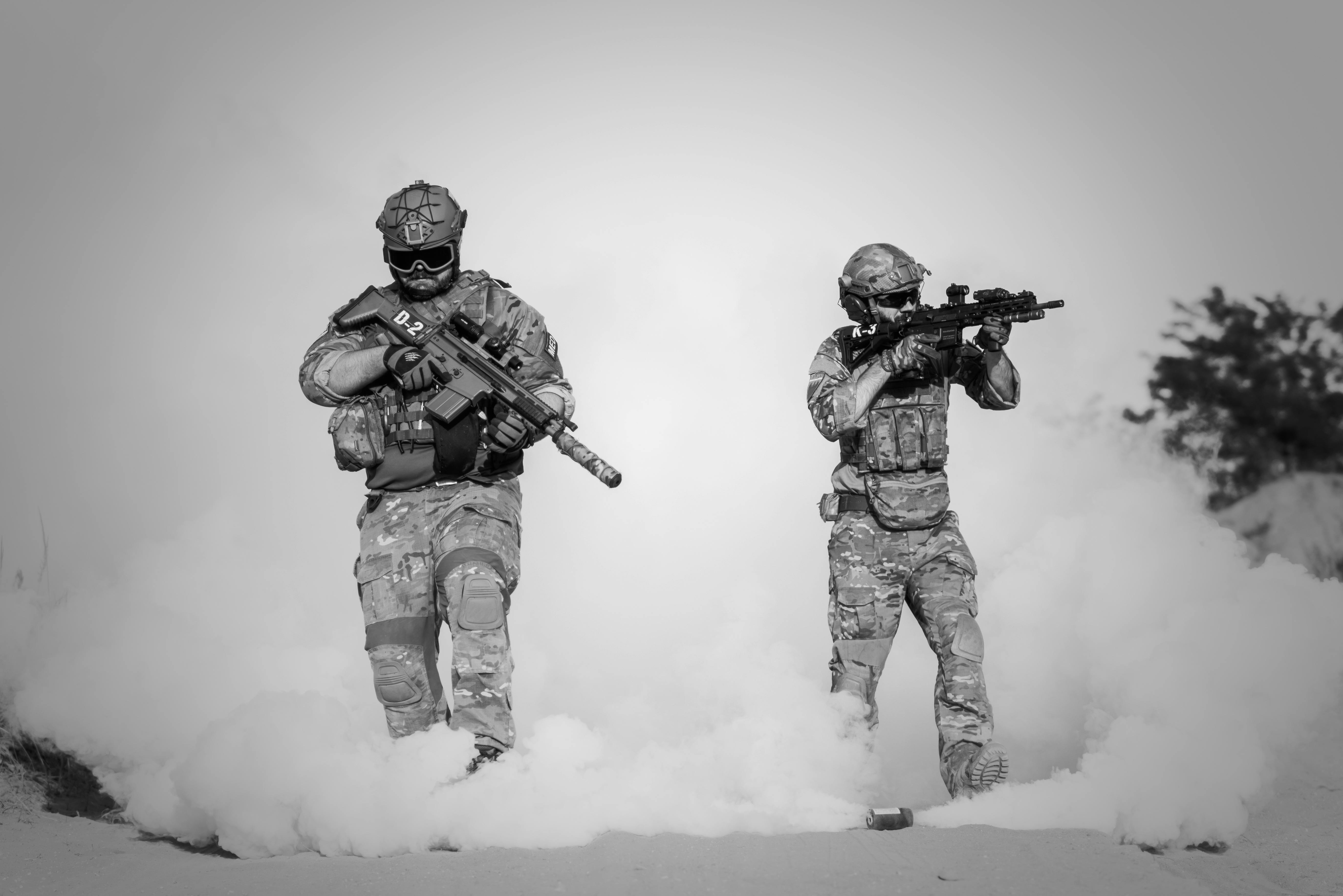 Black and white image of soldiers in a tactical operation amidst smoke and sand.