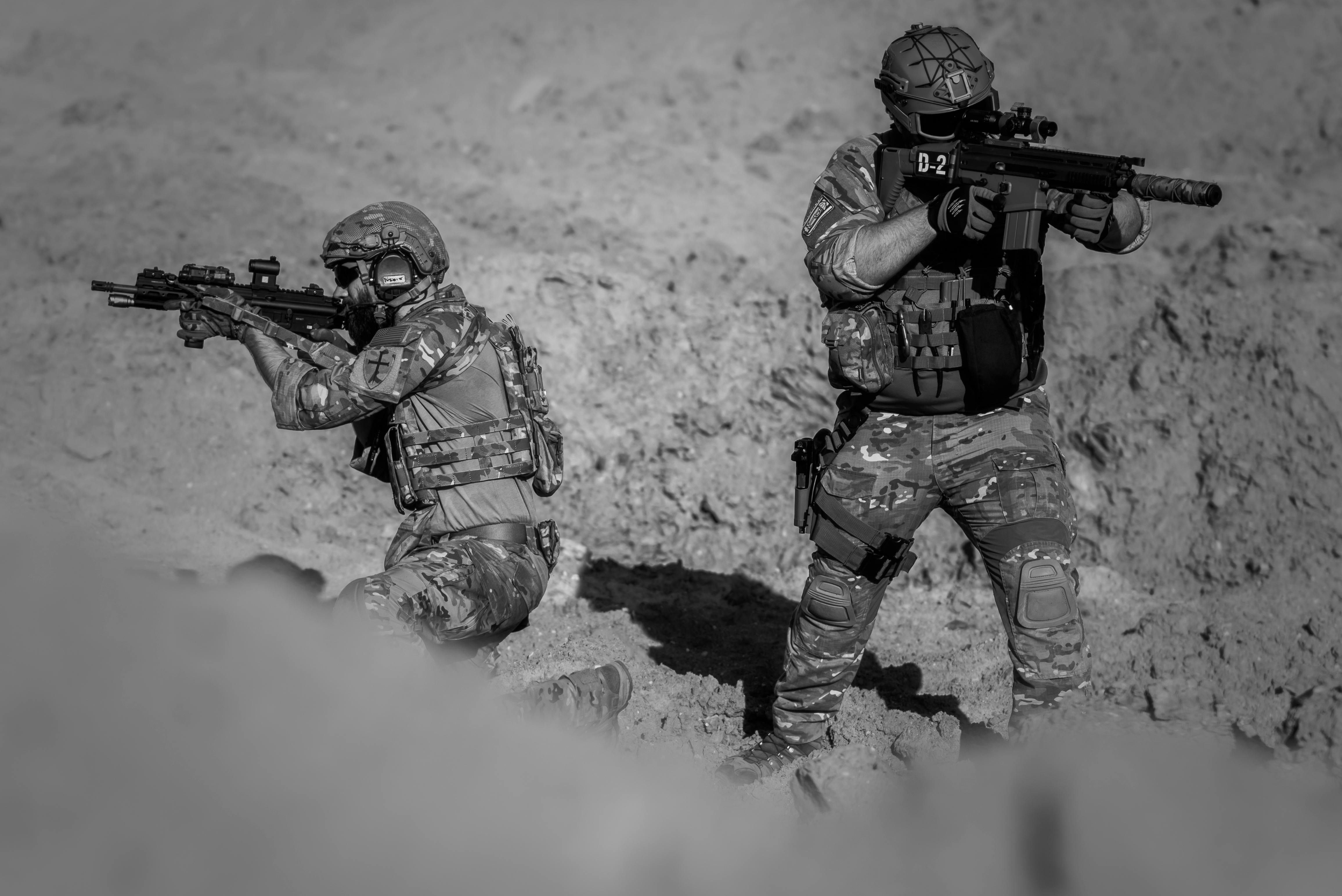 Two soldiers in camouflage and gear in a desert setting, engaged in a tactical action.